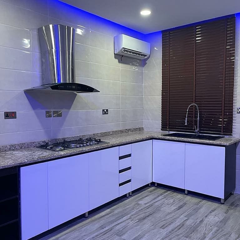 The modern kitchen features sleek white cabinetry complemented by granite countertops. A stainless steel hood and faucet highlight the space. Under-cabinet lighting casts a gentle glow, while dark wooden blinds provide control over natural light entering from the window.