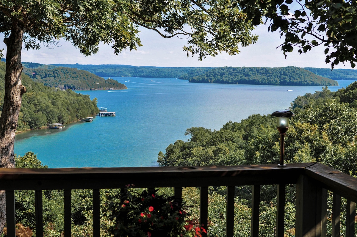 A serene view of Beaver Lake is captured from a wooden deck, surrounded by lush greenery. The water displays a gradient of blue hues, with distant hills visible under a clear sky. A flowering pot adds a touch of color to the railing.