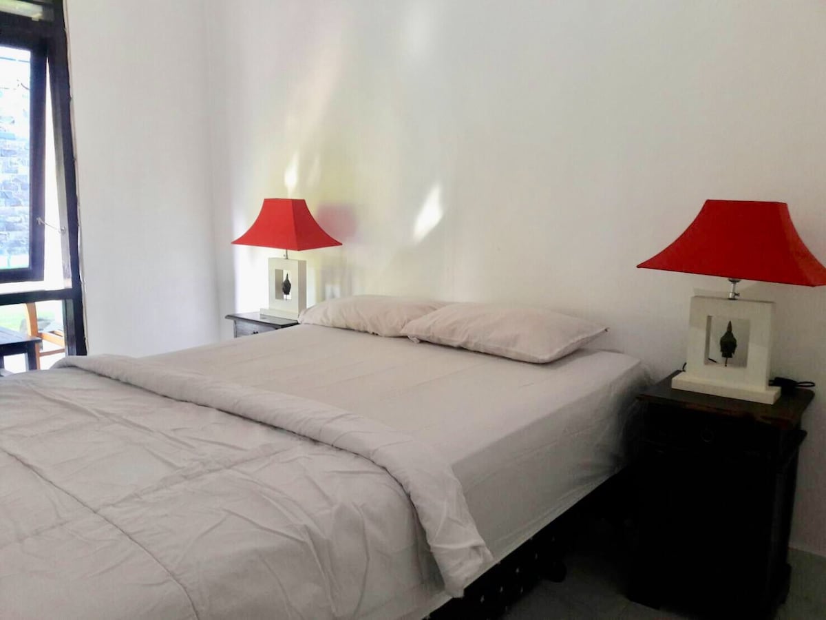 A bright bedroom showcases a spacious bed with neutral bedding. Each bedside table is adorned with a red lamp, providing a pop of color. Natural light filters through a nearby window, enhancing the simplicity of the white walls and soft furnishings.