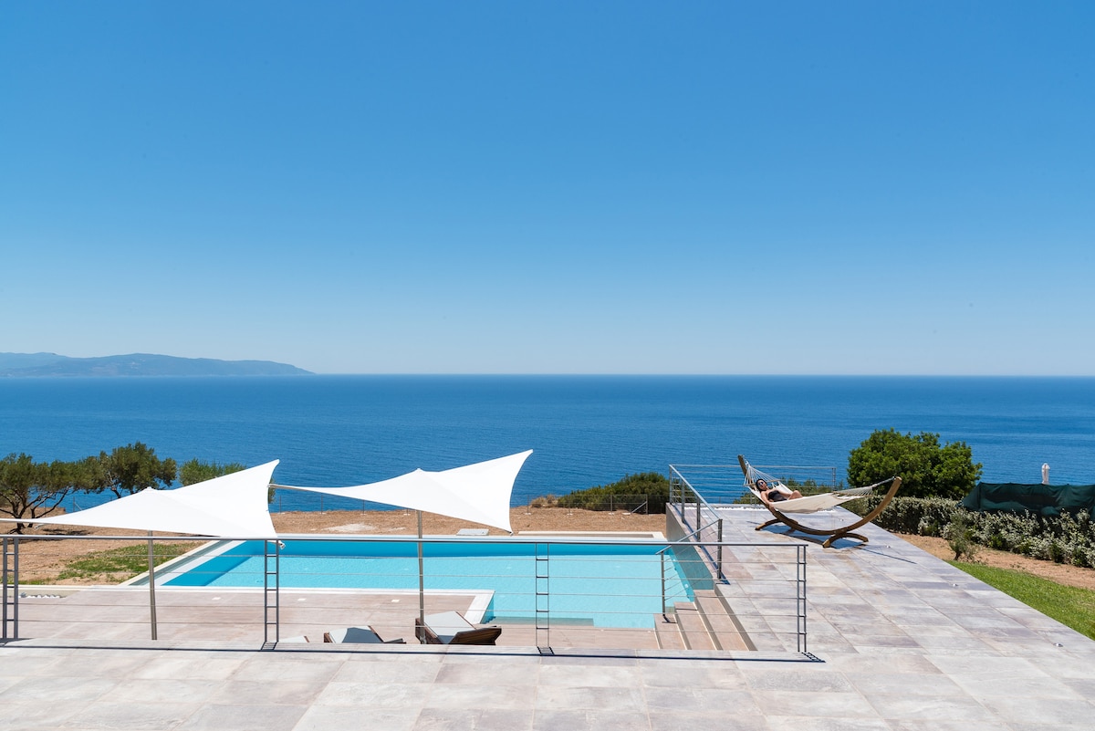 An infinity pool is situated at the edge of a spacious patio, with a clear view of the Ionian Sea in the background. Two white sunshades provide shade over the pool area, while a hammock is featured nearby under the bright blue sky.
