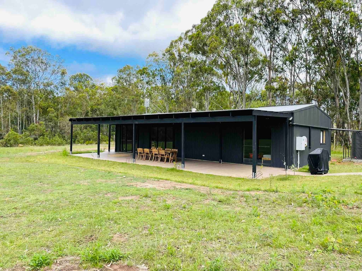 The converted barn features a spacious covered outdoor area with a long dining table, set against a backdrop of open grassy land and trees. Large windows provide ample natural light. The structure's sleek black exterior contrasts with the surrounding greenery.