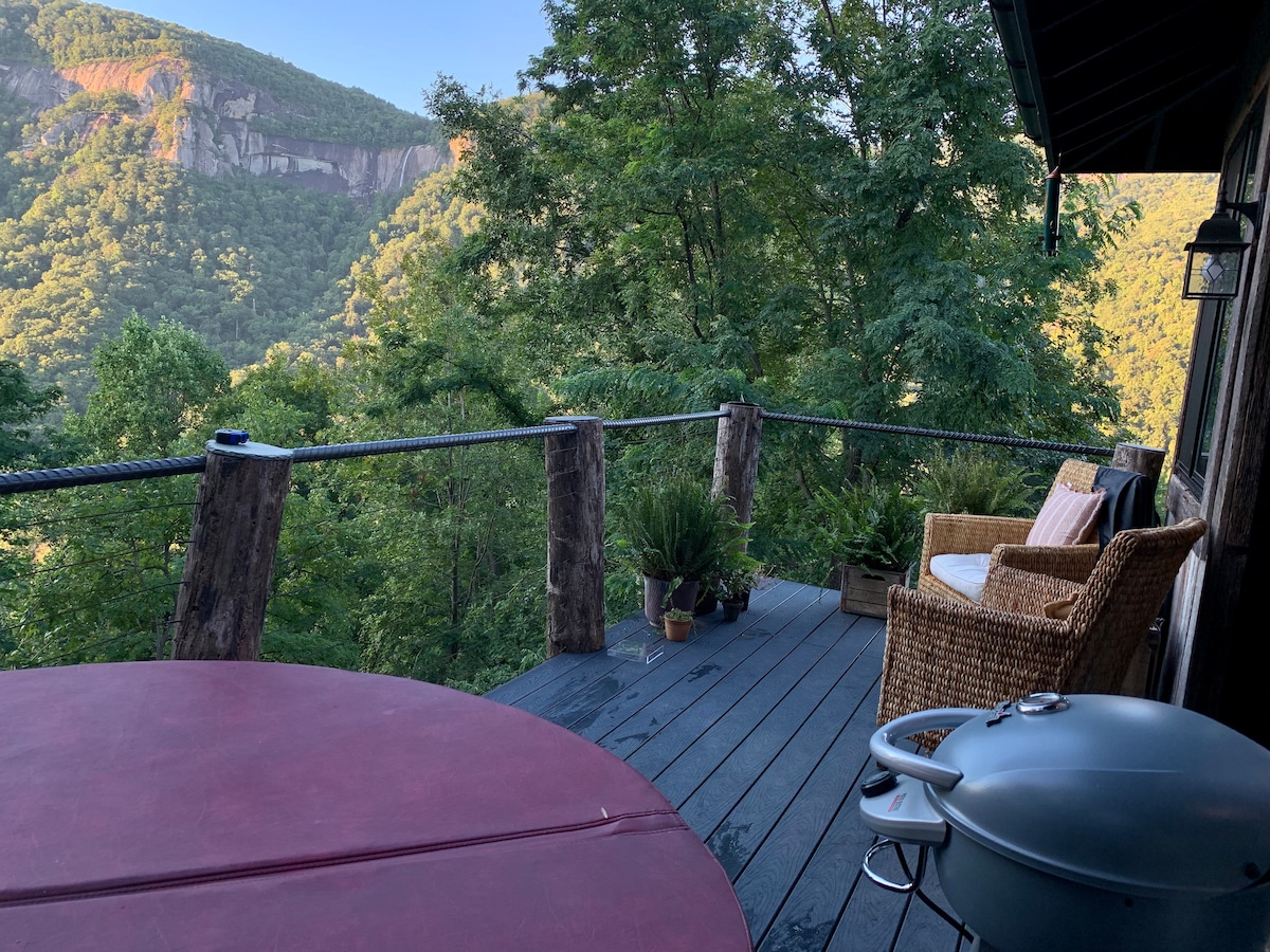 A spacious deck is shown with a hot tub and a grill, overlooking a lush mountain view. Comfortable seating made of woven material is positioned nearby, surrounded by greenery and decorative plants, creating an inviting outdoor space for relaxation.