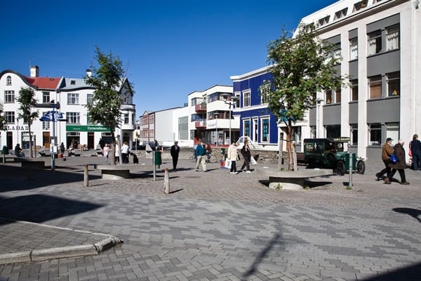 A bustling street scene showcases pedestrians walking on a paved square. Historic buildings are visible on either side, displaying a mix of architectural styles. Green trees line the square, providing shade, while blue skies and bright sunlight enhance the lively atmosphere.