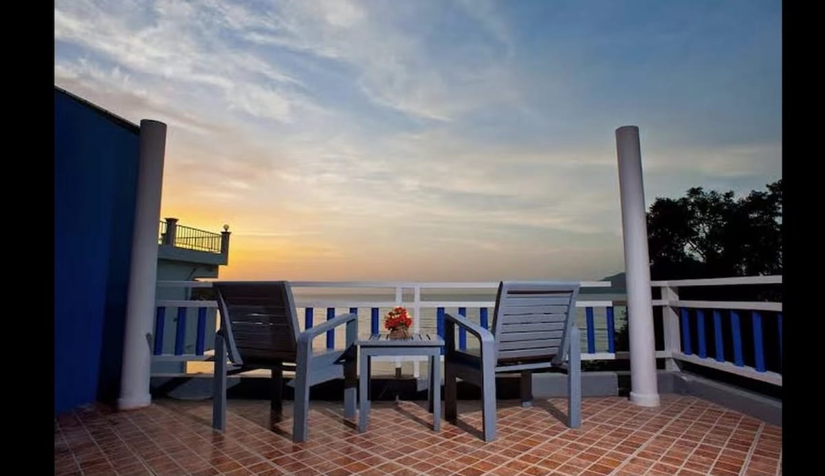 A terrace features two wooden chairs facing a small table, offering an unobstructed view of a sunset over the horizon. The space is framed by white railings and blue accents, with a tiled floor providing a warm backdrop for relaxation.