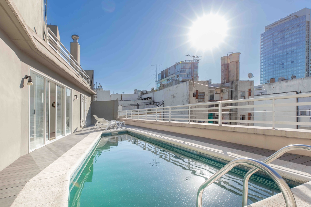 A rooftop swimming pool is featured, surrounded by a sunlit deck. Clear blue water reflects the sunlight, while lounge chairs are positioned nearby. The urban skyline provides a backdrop, with buildings visible in the distance under a clear sky.