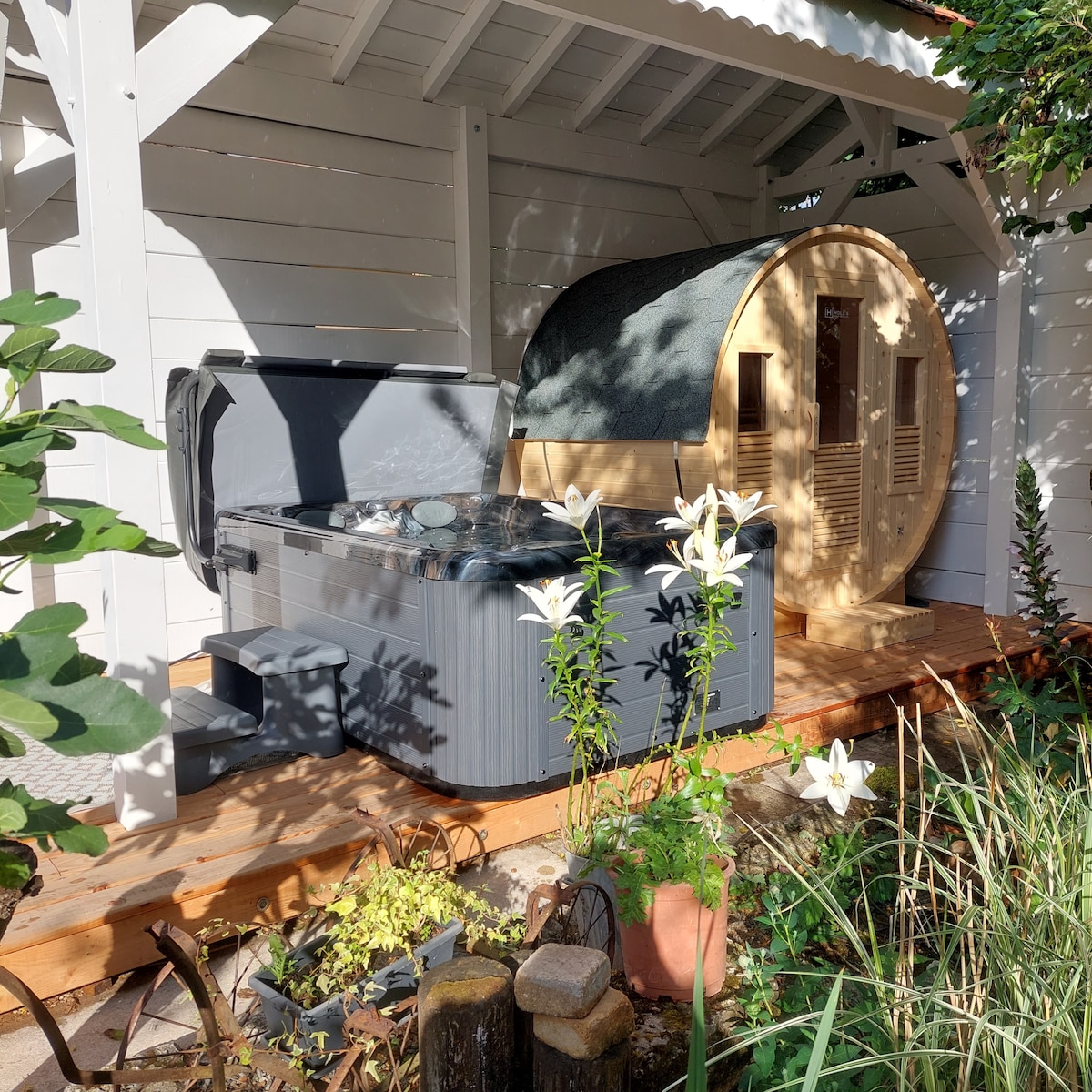 An outdoor spa area features a private hot tub alongside a wooden sauna cabin, both situated on a wooden deck. Lush greenery and blooming flowers surround the space, enhancing the tranquil setting. Natural light filters through, creating a calm atmosphere.