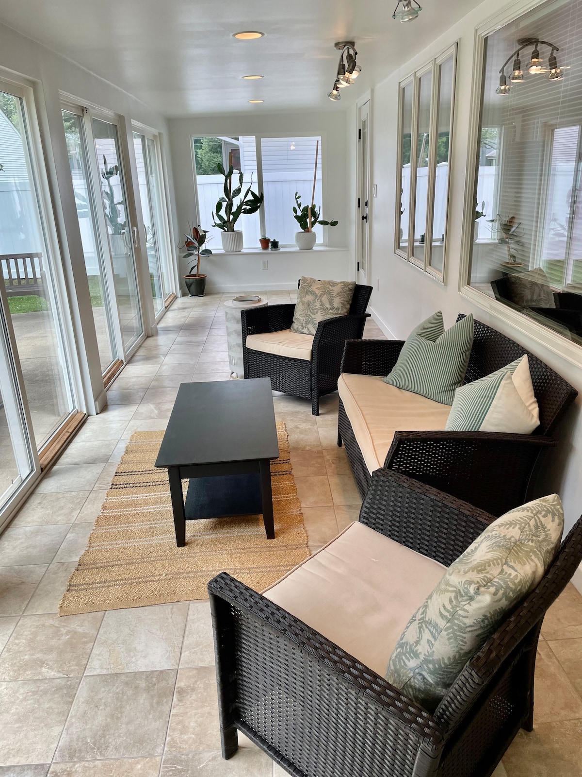 A sun porch features large windows allowing natural light to fill the space. Comfortable seating is arranged around a small black table, with potted plants positioned nearby. A light-colored rug adds softness to the tiled floor, enhancing the calming atmosphere.