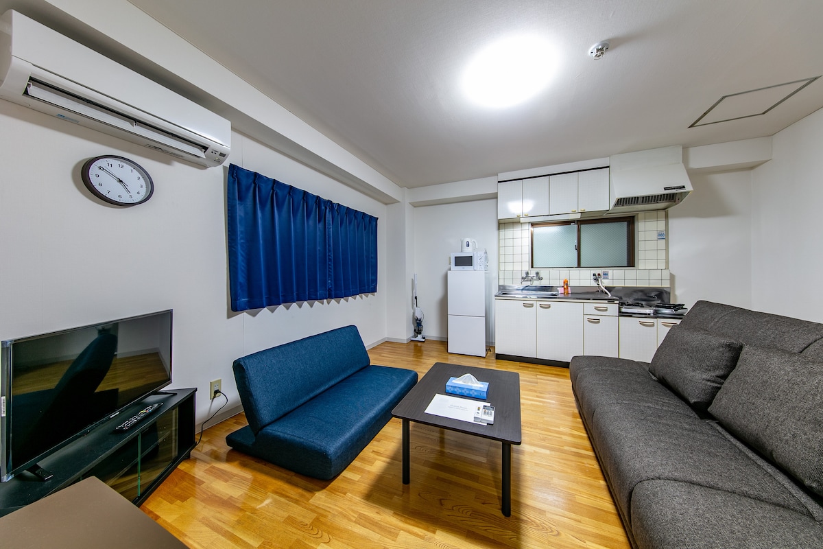 The living area features a grey sofa and a blue sofa placed opposite a low black coffee table, with a television set against the wall. A window is covered by dark blue curtains, while the kitchen area is equipped with white cabinets, a countertop, and essential appliances.