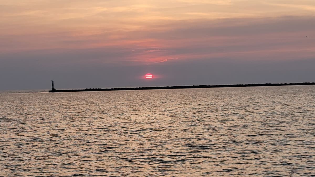 A serene sunset is captured over the water, with the sun dipping below the horizon. A distant lighthouse silhouette is visible on the rocky shoreline, creating a tranquil scene. The sky features soft hues of pink and purple, reflecting on the water's surface.