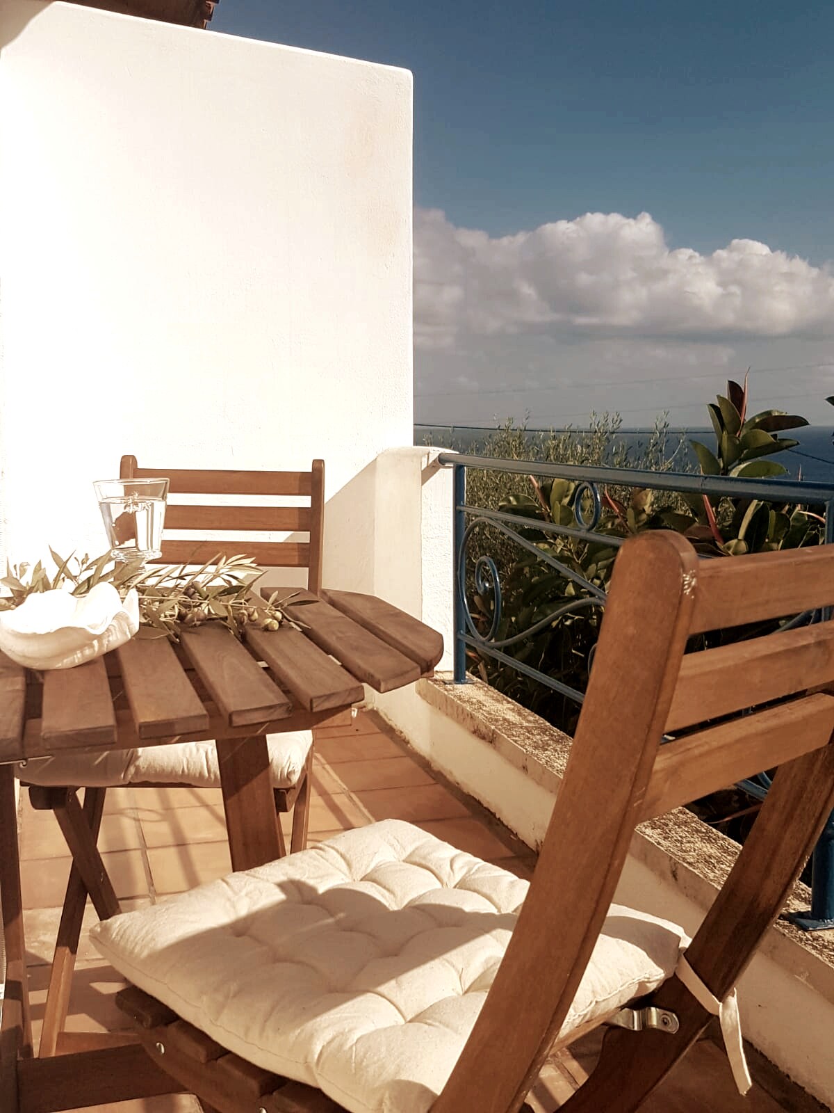 A wooden table and two chairs are arranged on a tiled balcony, providing a clear view of the sea and surrounding greenery. A glass of water is placed on the table, with soft cushions on the chairs for added comfort. The sky is partly cloudy.
