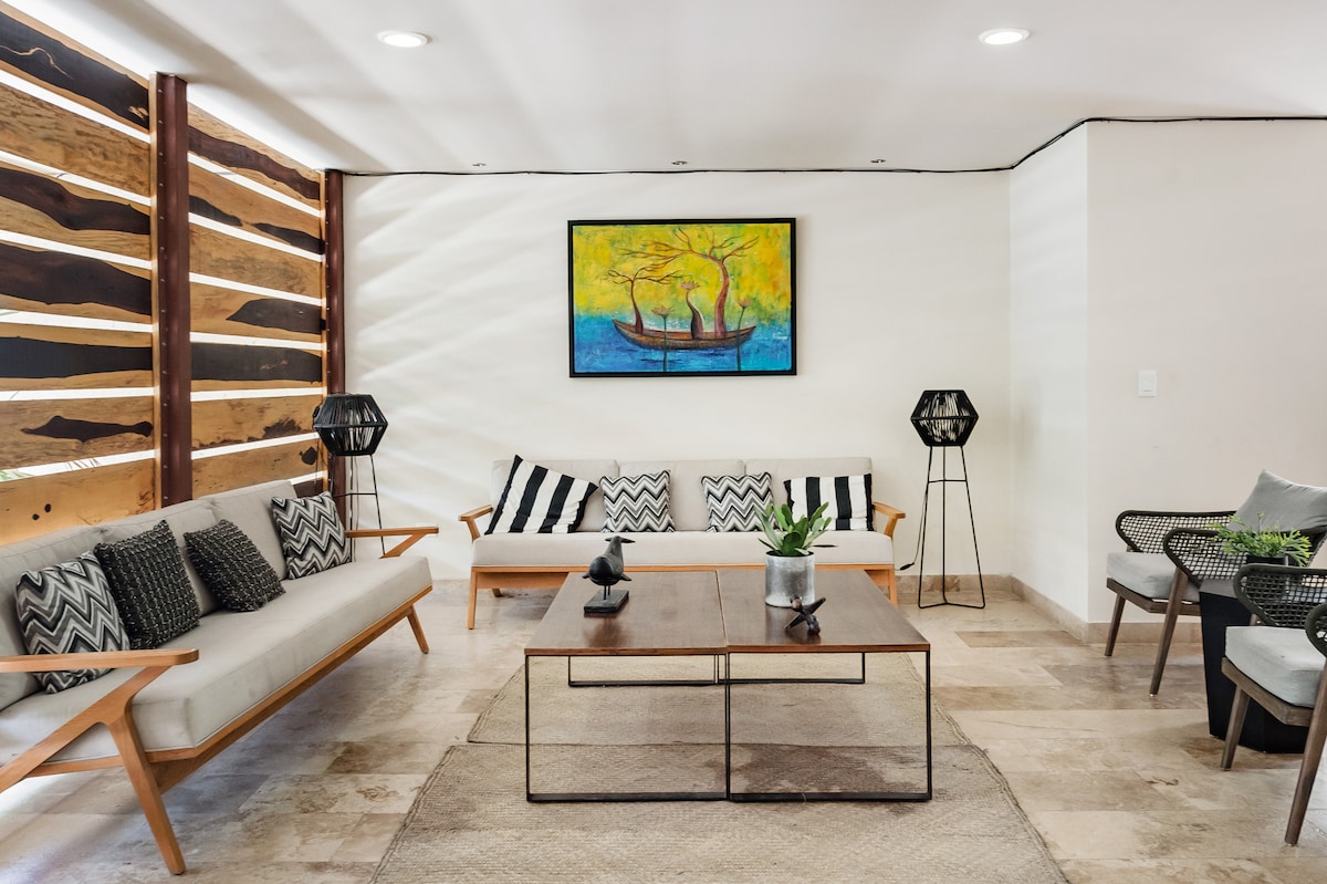 A modern living area features a neutral color palette with wooden accents. Two sofas are arranged facing a coffee table. A colorful painting hangs on the wall, complemented by natural light filtering through an artistic wooden partition.