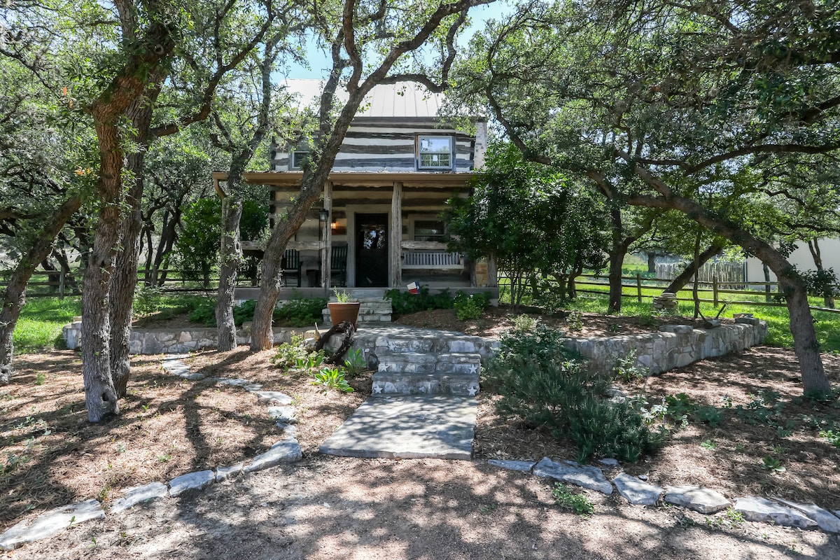 A rustic log cabin is nestled among trees, featuring a welcoming porch swing and rockers. A stone pathway leads to the entrance, surrounded by landscaped greenery. The two-story structure showcases classic architecture, inviting guests to relax in a serene outdoor setting.