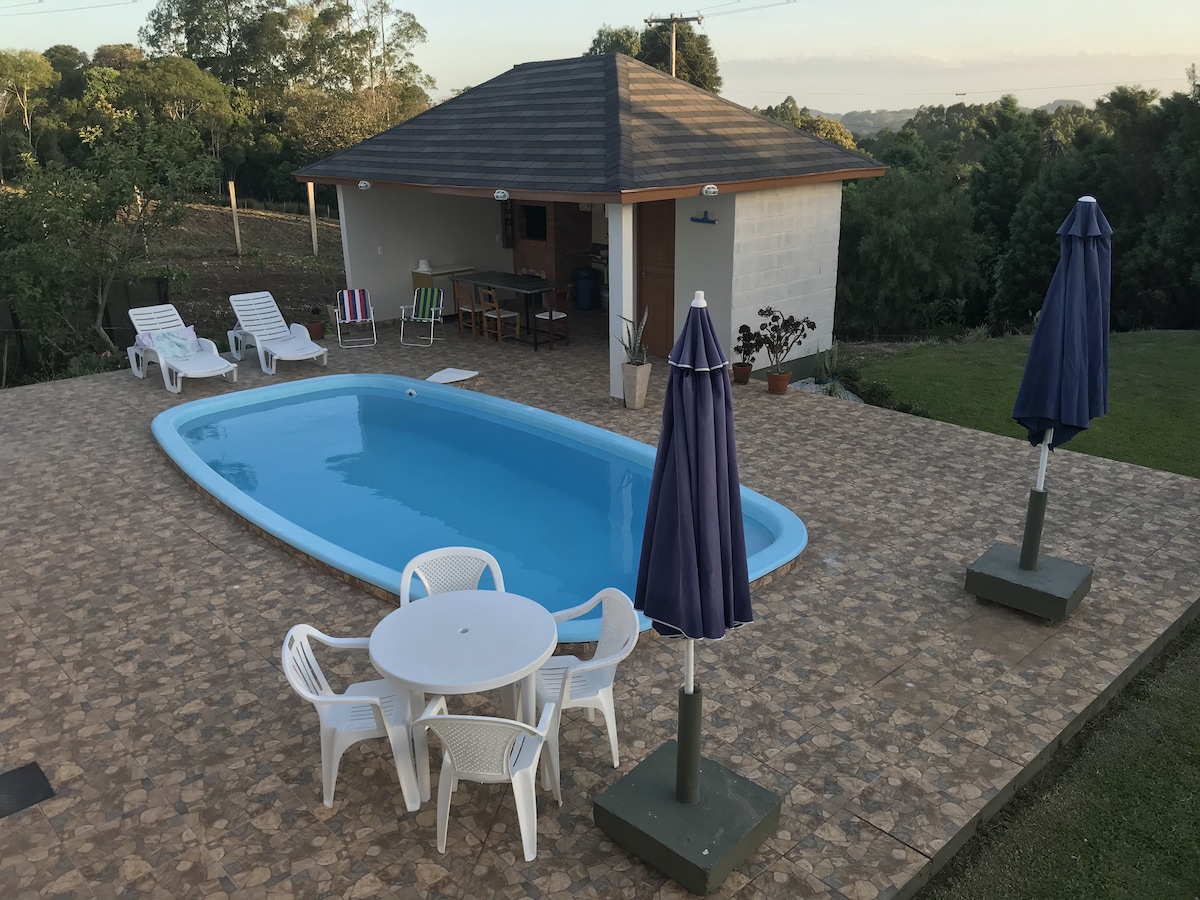 A pool surrounded by a spacious patio is featured, complemented by two umbrellas and a circular table with four chairs. The cozy outdoor kitchen area is visible in the background, set against a lush landscape of greenery.