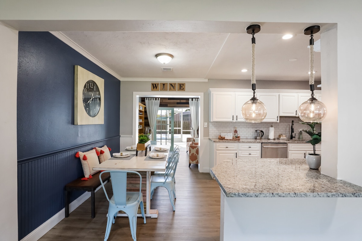 The dining area features a stylish table set for four, accompanied by elegant blue chairs. A wall clock adds character, while wooden accents enhance the space. The kitchen is visible, showcasing modern appliances and a granite countertop. Natural light enters through sliding doors, leading to the outdoor area.