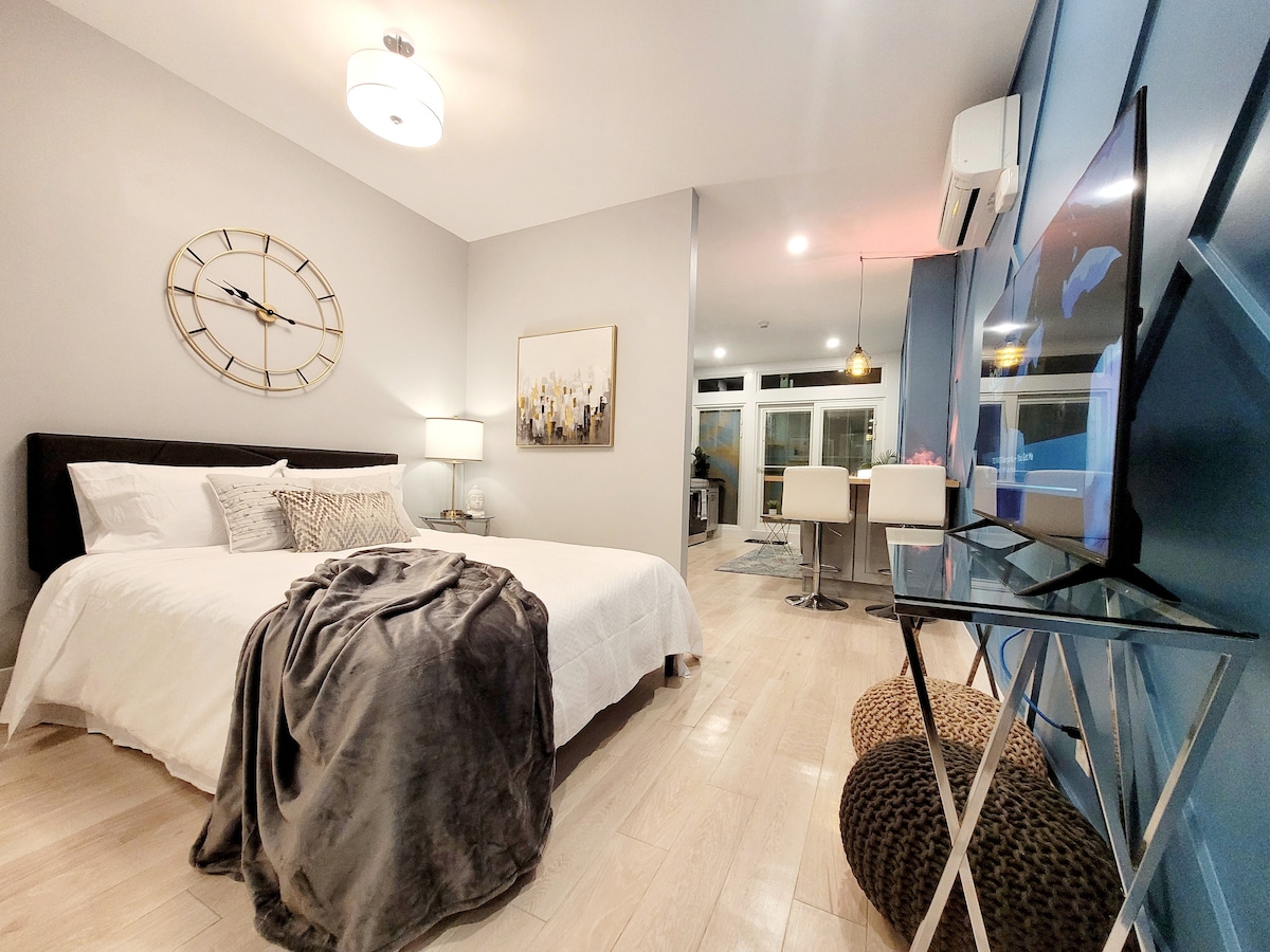 A bright and airy bedroom features a spacious bed dressed in soft linens, complemented by decorative pillows. A large wall clock adds a modern touch. Natural light fills the room through a sliding glass door, while a flat-screen TV is situated on a sleek stand.