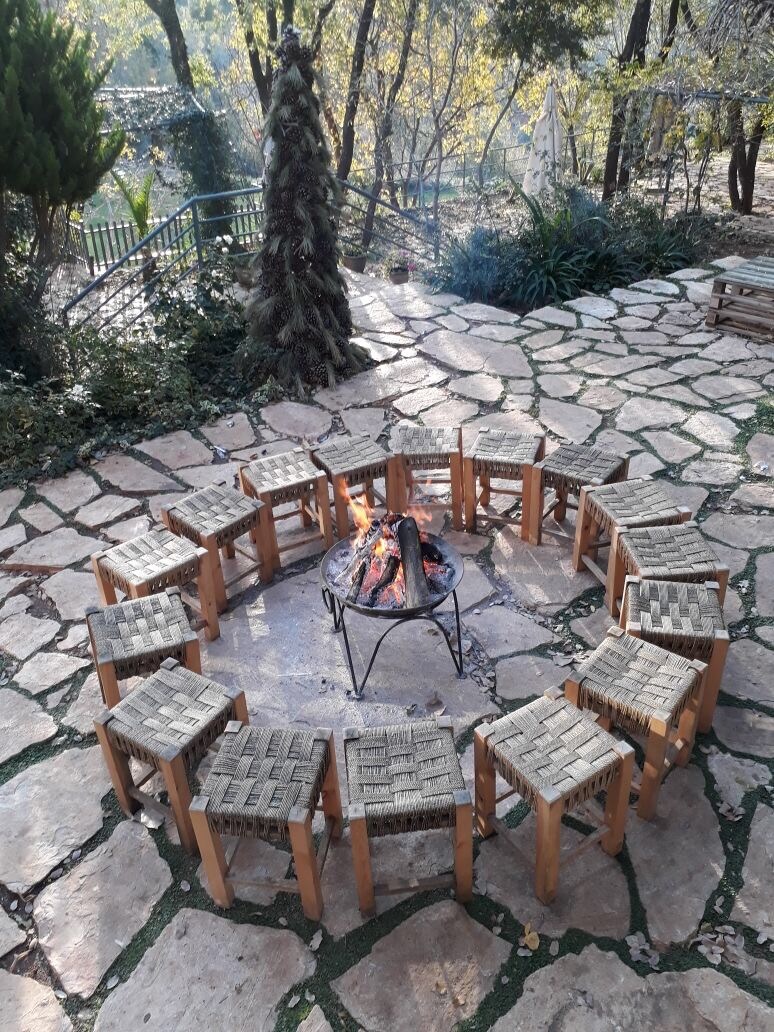 A circular arrangement of wooden stools surrounds a fire pit, situated on a stone patio. The area is bordered by lush greenery, with trees and plants visible in the background, creating a serene outdoor space for gathering.
