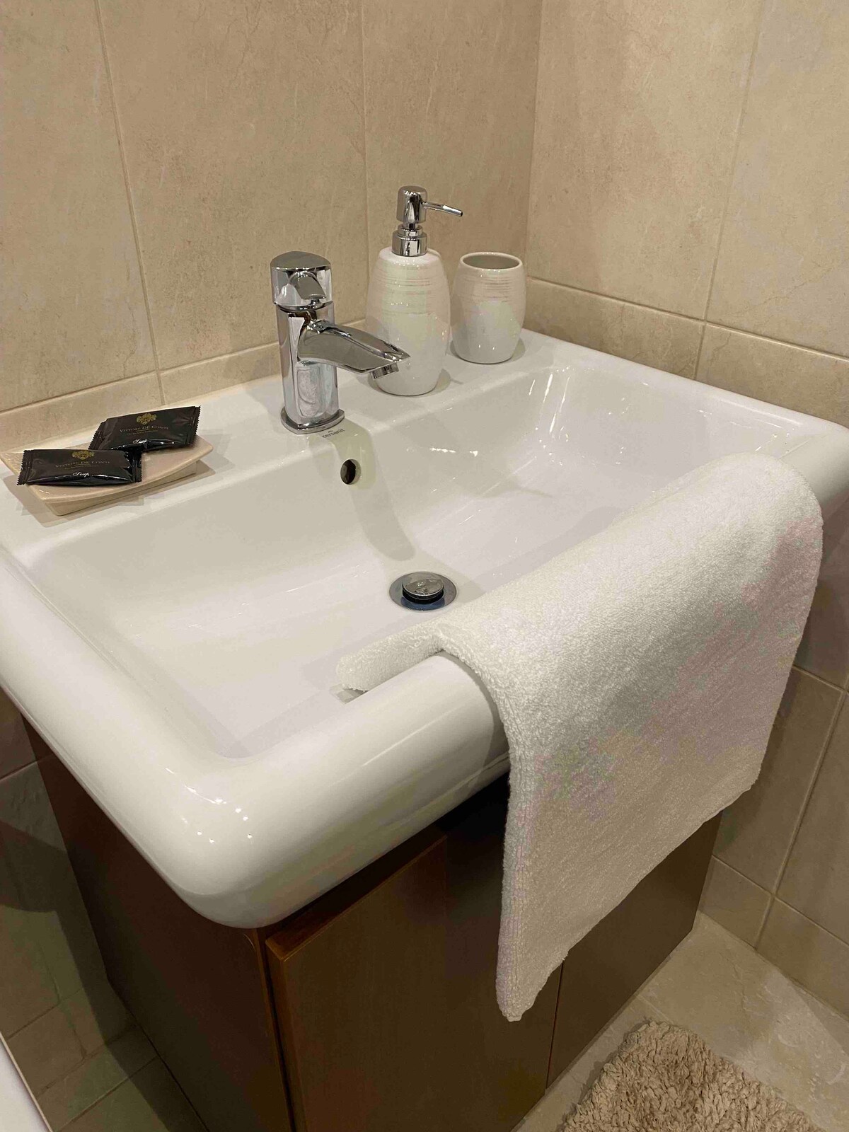 A clean sink is positioned in a modern bathroom, featuring a polished faucet and a soap dispenser. A neatly folded white towel rests on the edge of the sink. Small toiletries and a decorative cup are placed nearby, enhancing the functional design of the space.