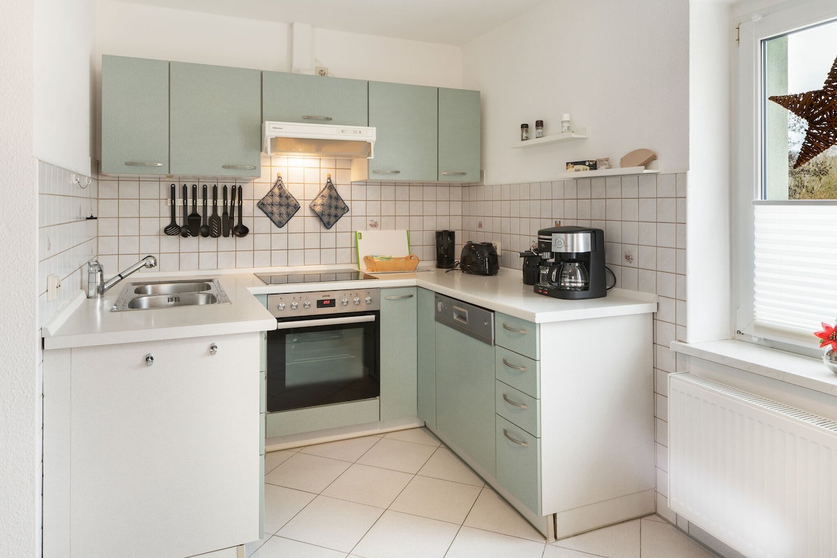 The kitchen is equipped with modern appliances, including a built-in oven and a coffee machine. Light-colored cabinets provide ample storage space, while a tiled backsplash complements the countertops. A window illuminates the area, showcasing a clean, organized environment for meal preparation.