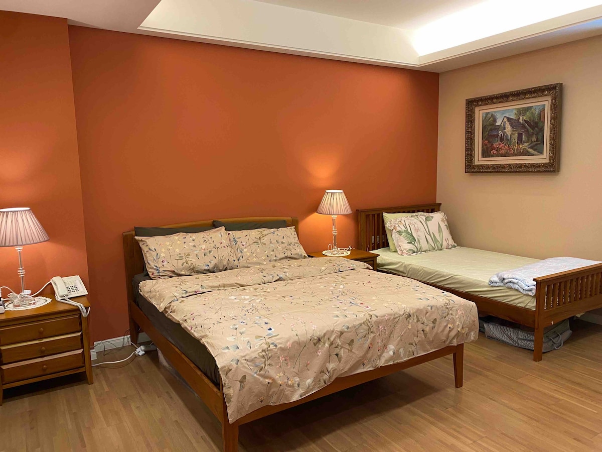 A spacious bedroom features two beds, one larger than the other, dressed in neutral-toned bedding with subtle patterns. Each bedside table includes a lamp, providing soft illumination. An accent wall in a warm orange hue complements the overall calming environment, while framed artwork adorns the adjacent wall.