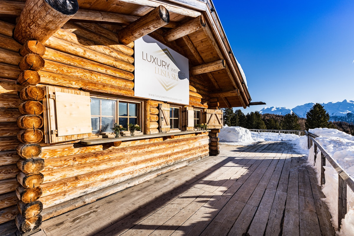A wooden chalet exterior is illustrated with log construction and large windows. A sign displaying 'LUXURY' is positioned centrally on the facade. A spacious wooden deck extends outward, covered with snow, with clear blue skies and distant mountain peaks visible in the background.