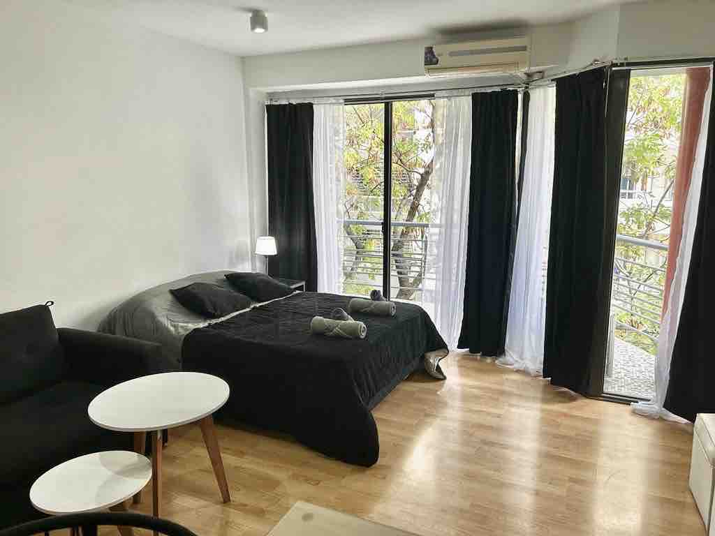A spacious studio apartment features a queen-sized bed with dark bedding and decorative pillows. A black sofa and modern coffee tables complement the hardwood floor. Floor-to-ceiling windows adorned with sheer and blackout curtains allow natural light and offer views of the greenery outside.