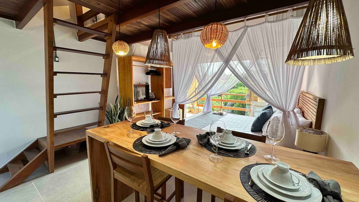 An inviting dining area features a wooden table set for four, complete with plates, cutlery, and wine glasses. Above, woven pendant lights provide warm illumination. A staircase leads to the upper level, while sheer drapes frame the opening to the outdoor space beyond.