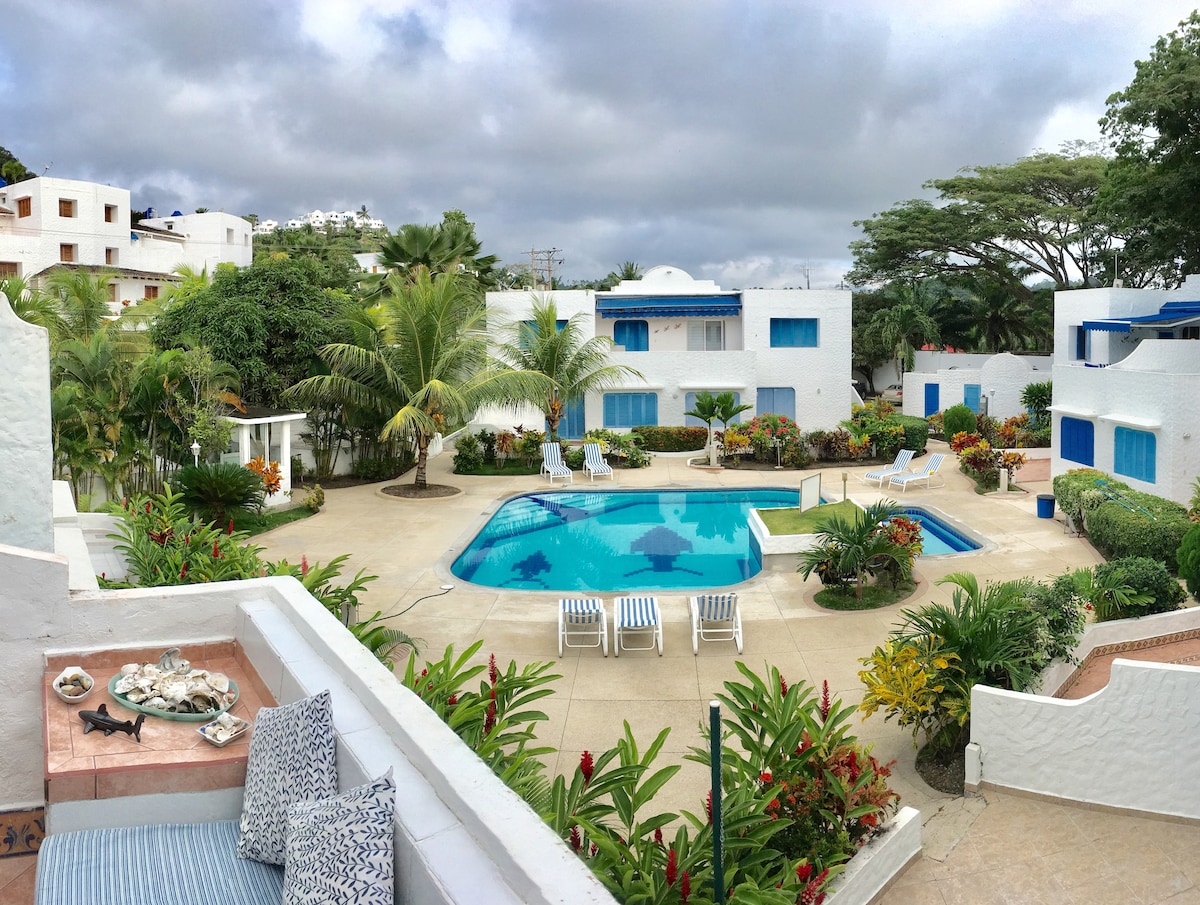 A serene outdoor area is featured, showcasing a swimming pool surrounded by lush gardens and palm trees. Sun loungers are positioned around the pool, and colorful flowers complement the tropical setting. The buildings, painted in white, reflect a relaxed architectural style.