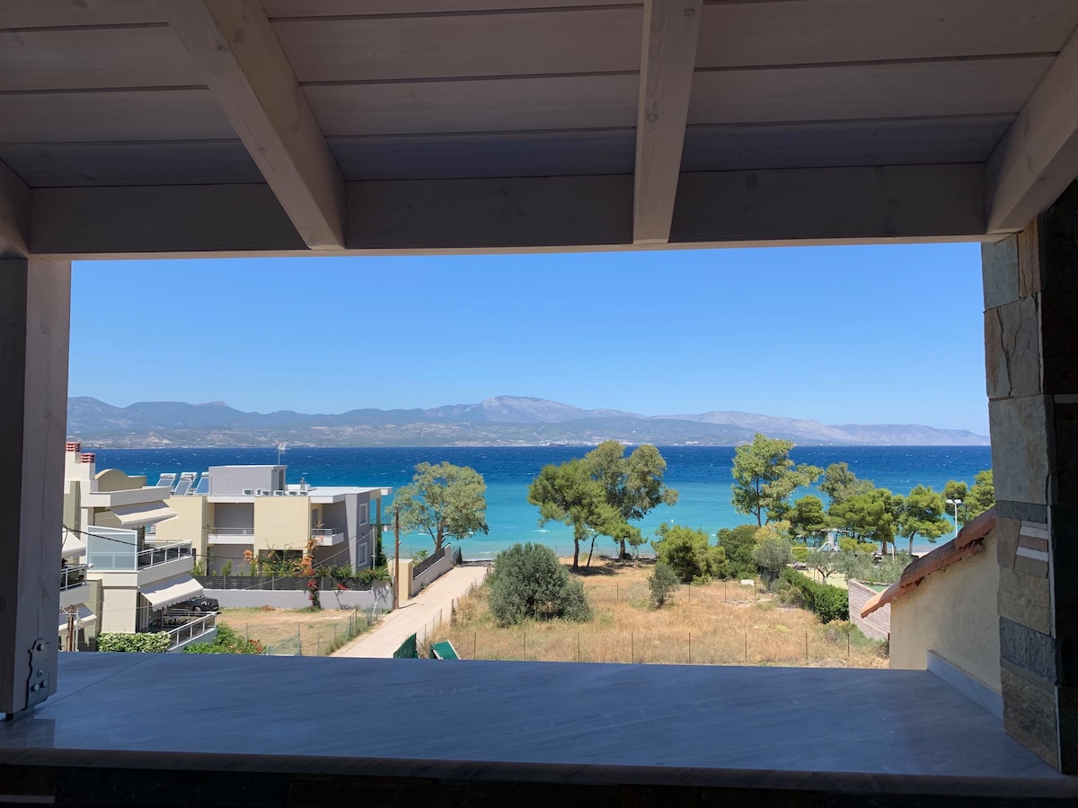A view of the Argosaronic Gulf is framed by an open balcony, showcasing the vibrant blue water and greenery of nearby trees. The horizon features undulating hills under a clear blue sky, creating a serene backdrop for relaxation.