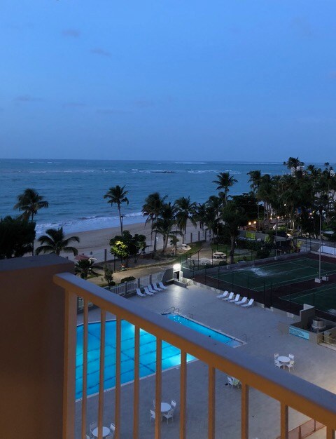A serene evening view reveals the ocean, gently lapping against the shore. The illuminated swimming pool reflects a soft glow, surrounded by palm trees. A tennis court can be seen in the distance, with lounge chairs positioned for relaxation. The sky shifts from light to dark as sunset approaches.