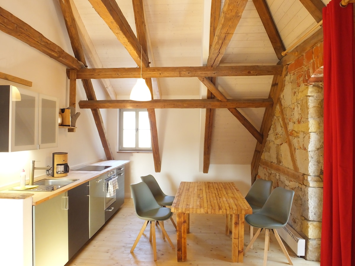 The living area features exposed wooden beams and a rustic stone wall. A dining table with six chairs is positioned near the kitchen, which includes modern appliances. A pendant light hangs above the table, and a large window allows natural light to fill the space.