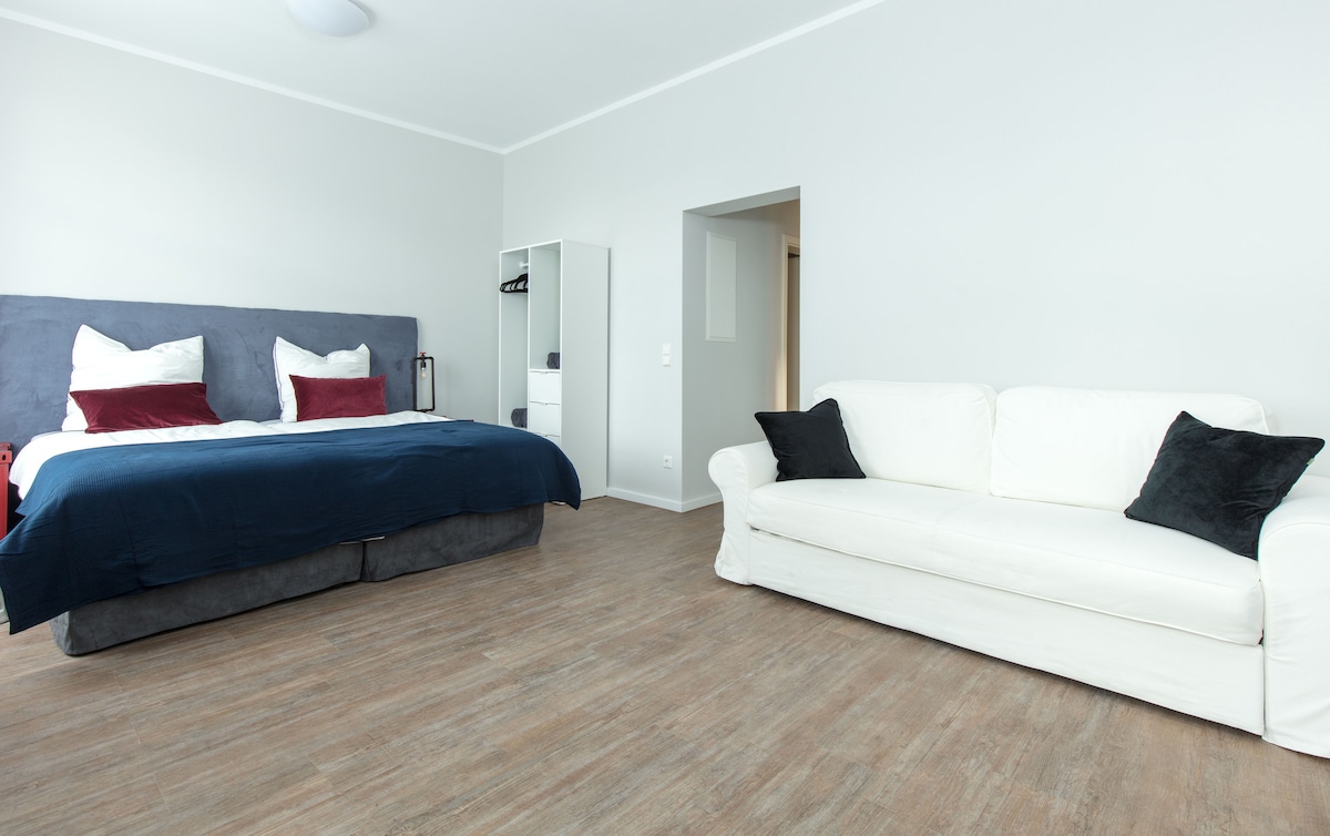 A spacious room is presented with a large bed featuring dark blue bedding and white pillows, accompanied by a cozy white sofa with two dark cushions. Light wooden flooring creates a warm atmosphere, complemented by neutral-colored walls.