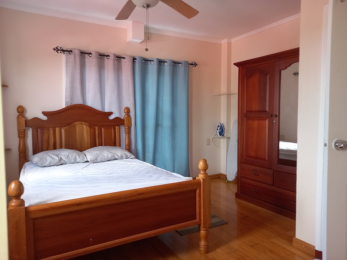 A warm bedroom features a wooden bed with a light-colored bedspread, complemented by two pillows. A ceiling fan is present above, and soft curtains frame a window. A wooden wardrobe is positioned against the wall, and an iron is neatly placed nearby.