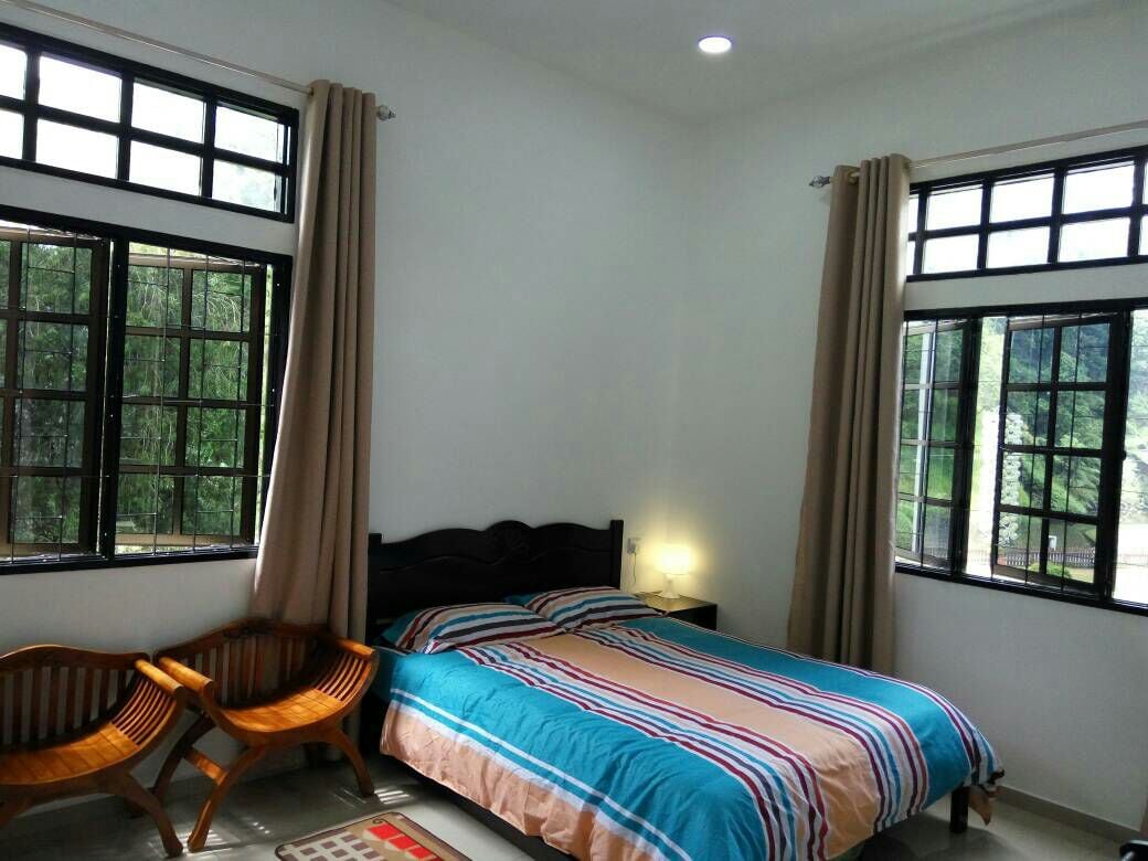 A well-lit master bedroom features a spacious bed covered with striped bedding. Two large windows are adorned with neutral curtains, allowing natural light to flow in. Two wooden chairs are positioned beside the bed, creating a simple yet functional seating area.