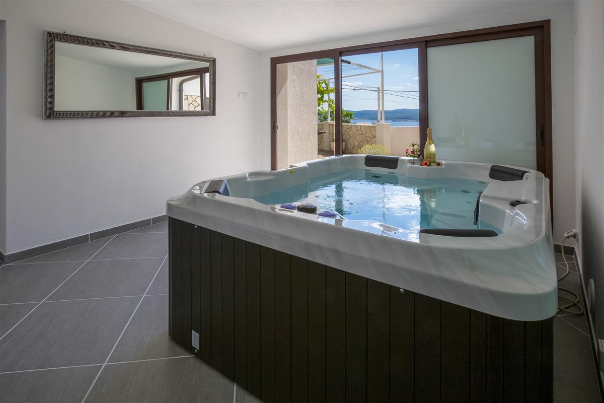 A whirlpool is positioned in a bright, spacious room, offering a view of the sea through large glass doors. The sleek design features a dark wooden base, with various seating areas along the edge. Sunlight reflects off the surface of the water, enhancing the calming atmosphere.