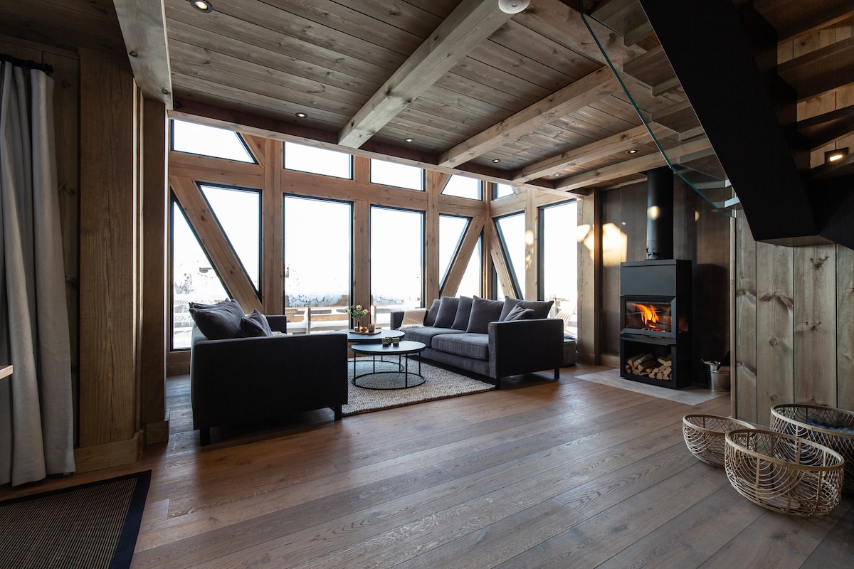 A spacious living area features large windows showcasing mountain views. A modern sofa set is arranged around a coffee table, positioned atop a woven rug. A wood-burning stove adds warmth to the room, complemented by natural wooden walls and flooring.