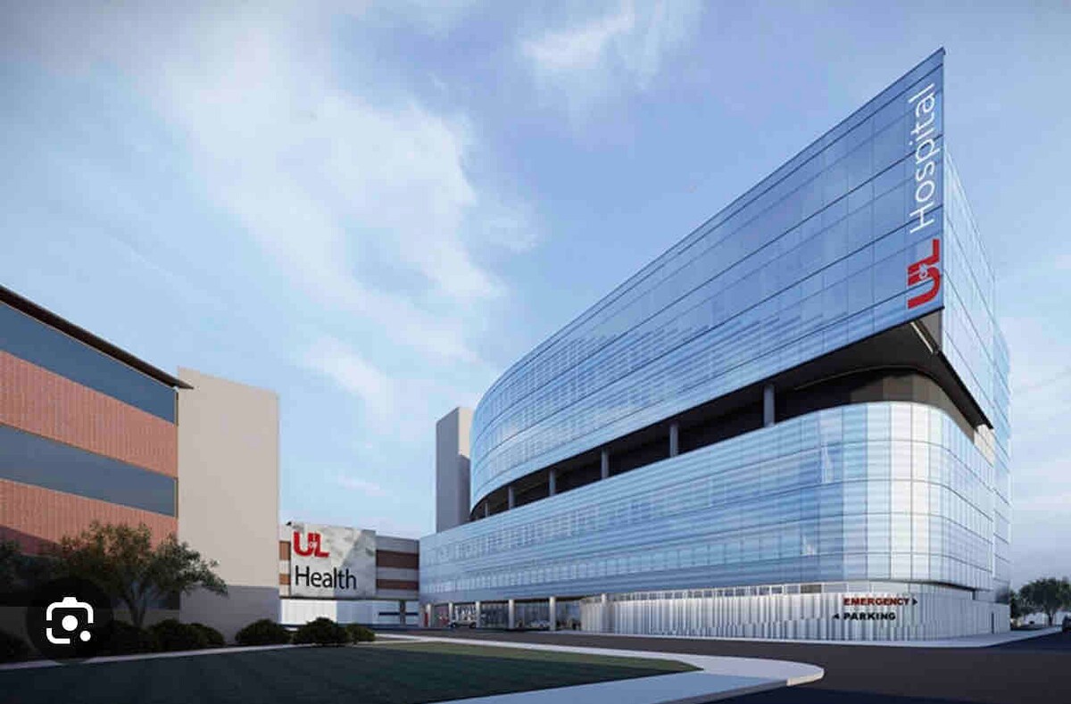 A modern hospital building is presented, featuring a sleek glass facade and a curved design. The prominent 'UL Health' logo is displayed clearly on the top, alongside emergency and parking signs. Surrounding green landscaping adds a touch of nature to the urban setting.