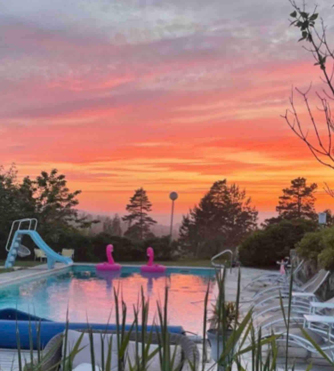 A serene outdoor pool area is presented, featuring a water slide and two pink floatation devices. Lush greenery surrounds the pool, and a vibrant sunset sky casts warm hues over the scene, creating a tranquil atmosphere. Several lounge chairs are positioned nearby.