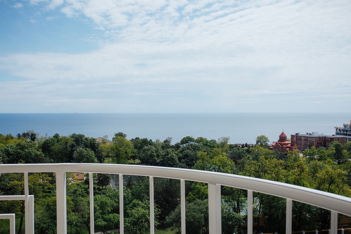A view from a balcony showcases a wide expanse of the sea merging with the sky, framed by lush greenery of trees below. The open horizon offers a sense of tranquility and connection to nature, contributing to a calm atmosphere.