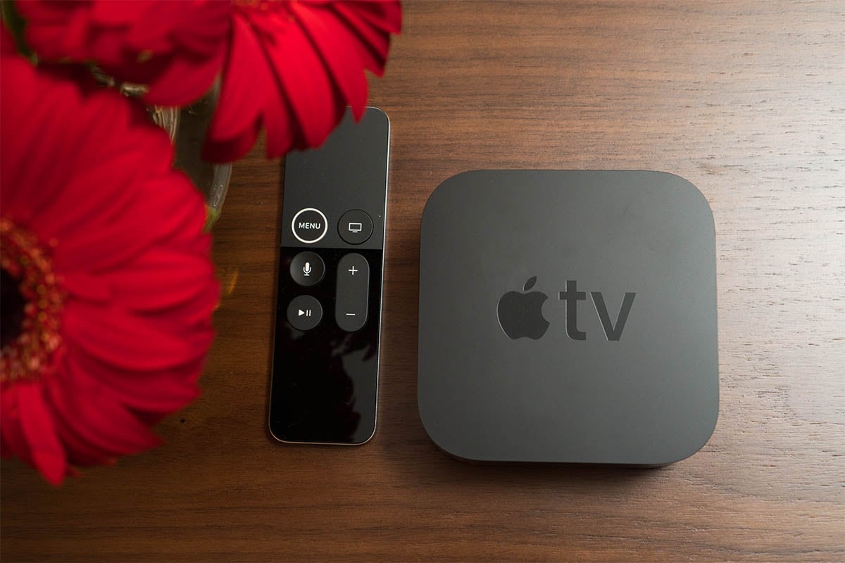 An Apple TV device is positioned next to a remote control on a wooden surface. A vase of vibrant red flowers is visible in the background, adding a touch of color to the scene.