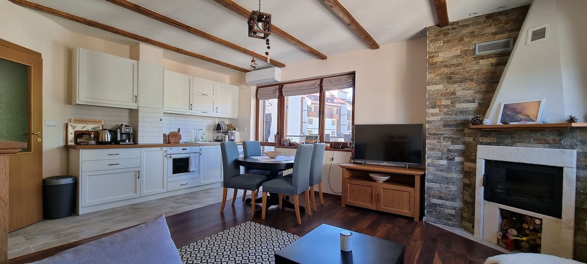 A cozy living space features a modern kitchenette and a dining area with a table and four chairs. A comfortable seating arrangement is complemented by a flat-screen TV. Natural light enters through large windows, and a stone fireplace adds warmth to the ambiance.