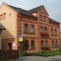 A brick building with a warm-toned facade features three architectural sections, highlighted by distinctive windows and decorative elements. Flower boxes adorn some windows, while a welcoming entrance is visible in the foreground. The structure is set in a peaceful setting.