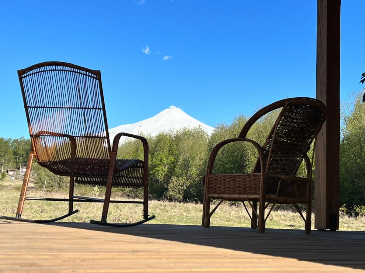 Two textured seating chairs are positioned on a wooden deck, overlooking a vast green landscape. In the background, a snow-capped volcano is visible against a clear blue sky. The scene captures a tranquil moment in nature.