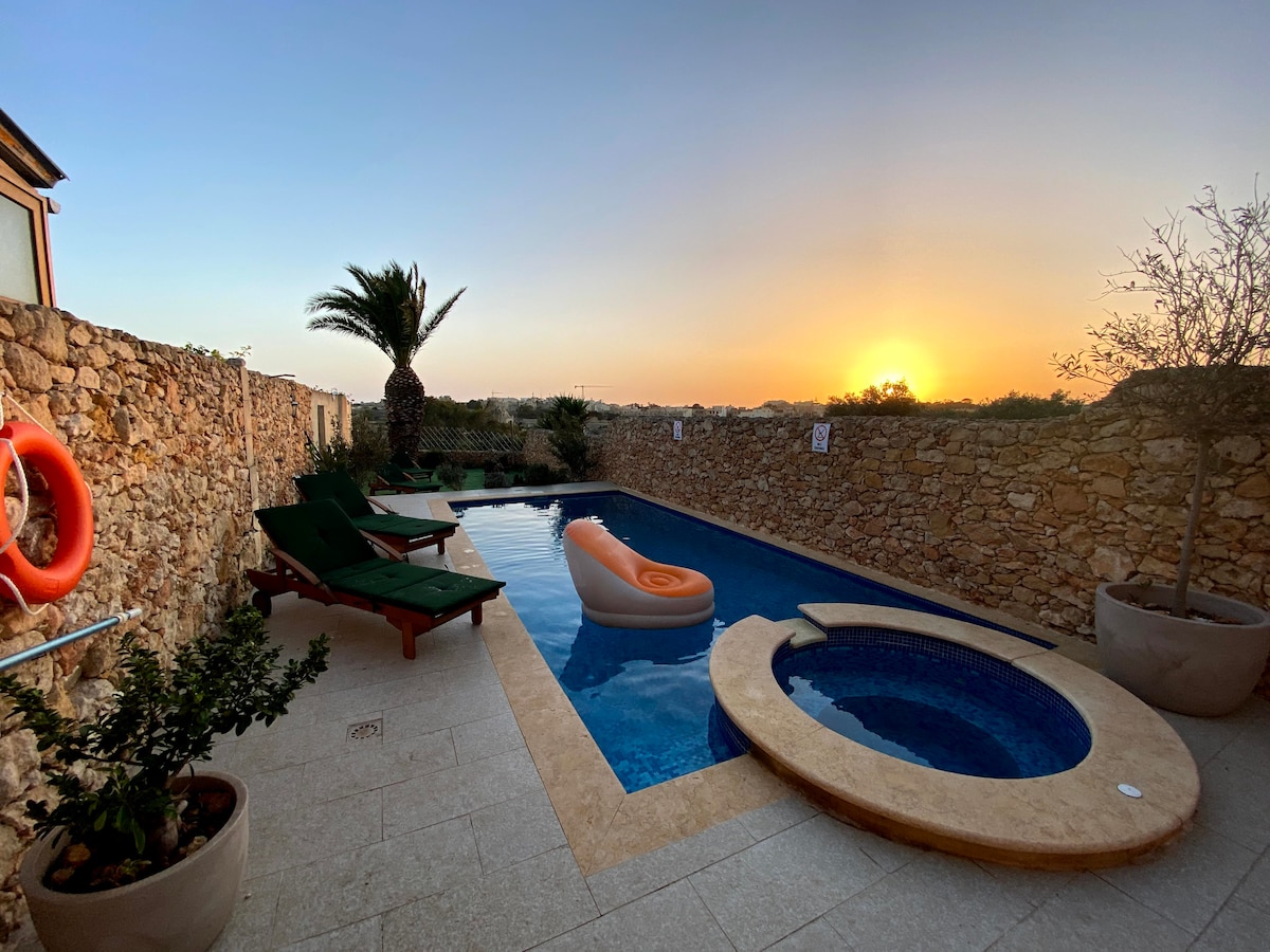 A private pool area is framed by natural stone walls, with lounge chairs positioned nearby. An inflatable float rests in the water, and a small hot tub is integrated into the pool design. Palm trees are visible in the background, illuminated by the sunset.