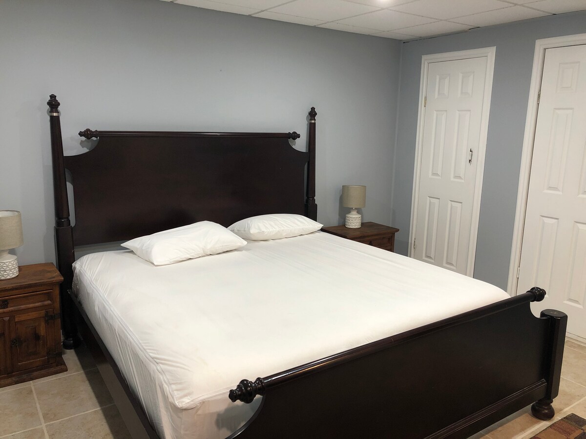 A spacious bedroom is featured, furnished with a dark wood bed frame and two nightstands with lamps. The room has light grey walls and is designed with a neutral aesthetic. Two doors are visible, leading to additional areas of the suite.