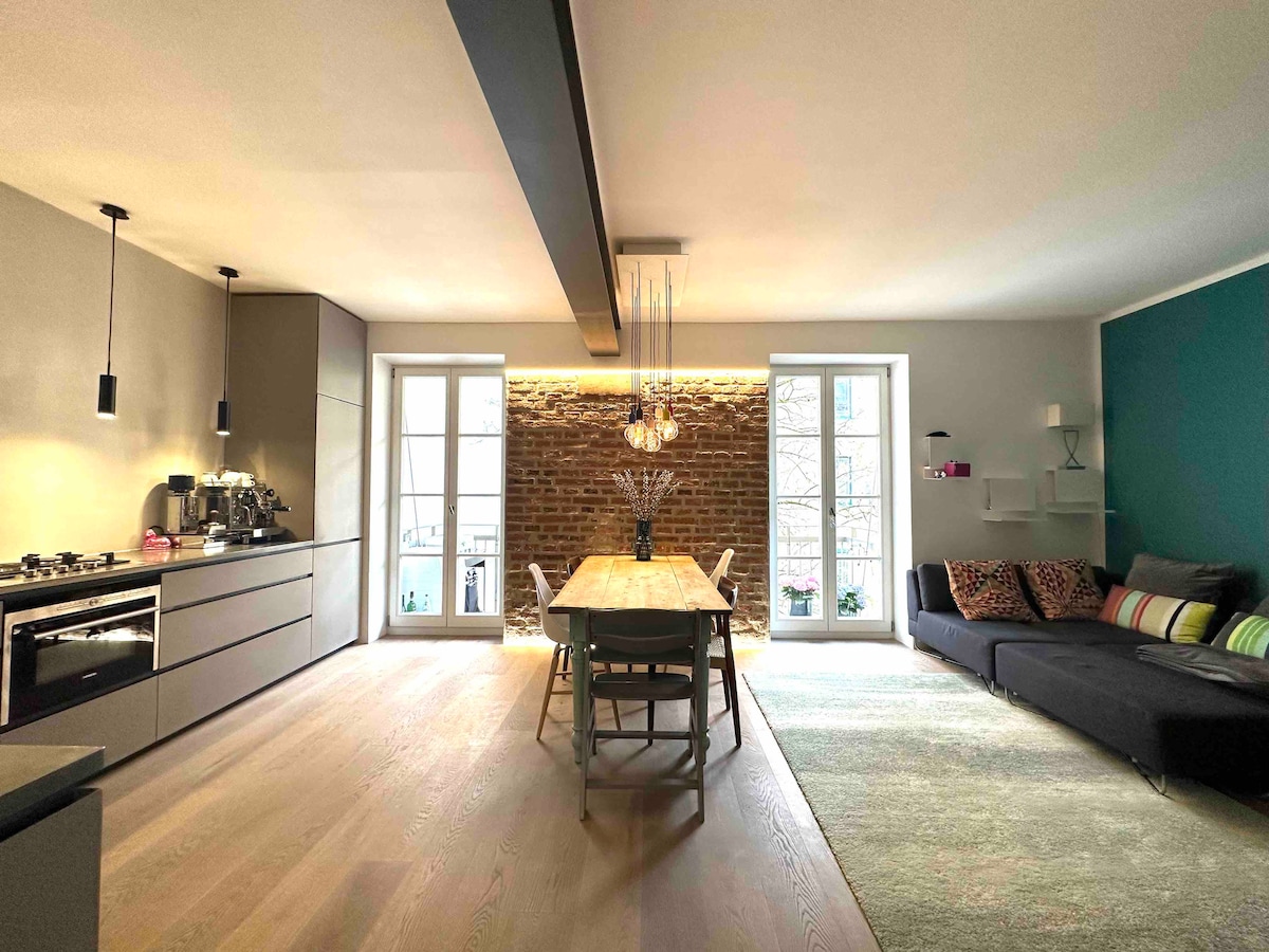 An open kitchen is paired with a dining area featuring a wooden table surrounded by chairs. A cozy sofa with colorful cushions is situated next to a green accent wall. Large windows allow natural light, highlighting the exposed brick wall.