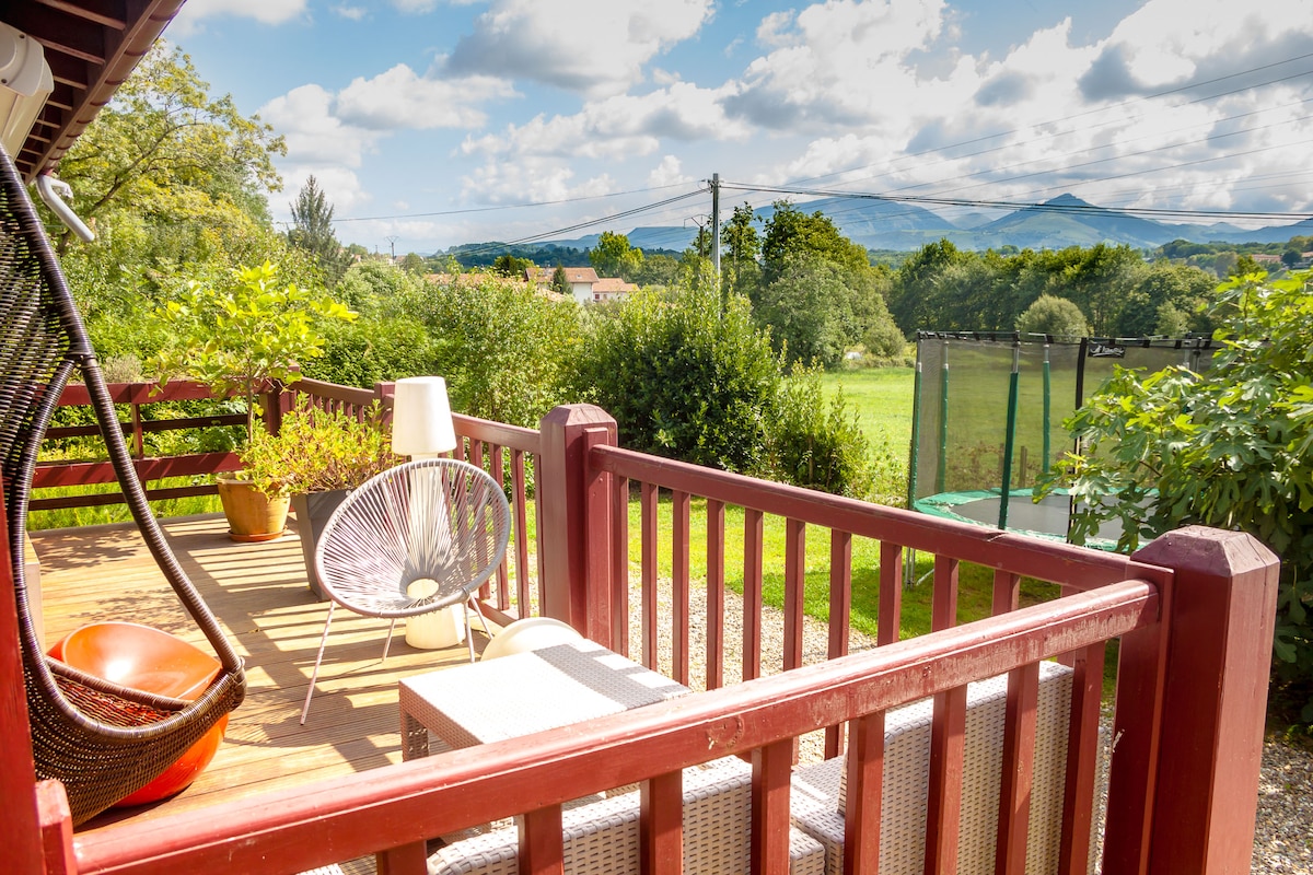 An outdoor deck features comfortable seating arrangements surrounded by lush greenery. Scenic views of mountains are visible in the background, adding to the serene environment. A hanging chair and potted plants contribute to a relaxed outdoor atmosphere.