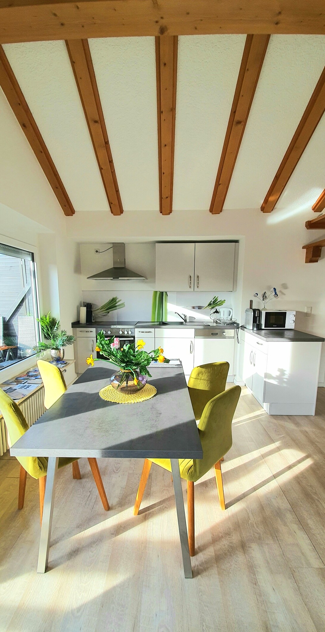 A bright kitchen area is designed with wooden beams overhead. A dining table with four chairs sits beside a sunny window. The kitchen includes modern appliances and ample counter space, creating a functional atmosphere for preparing meals.