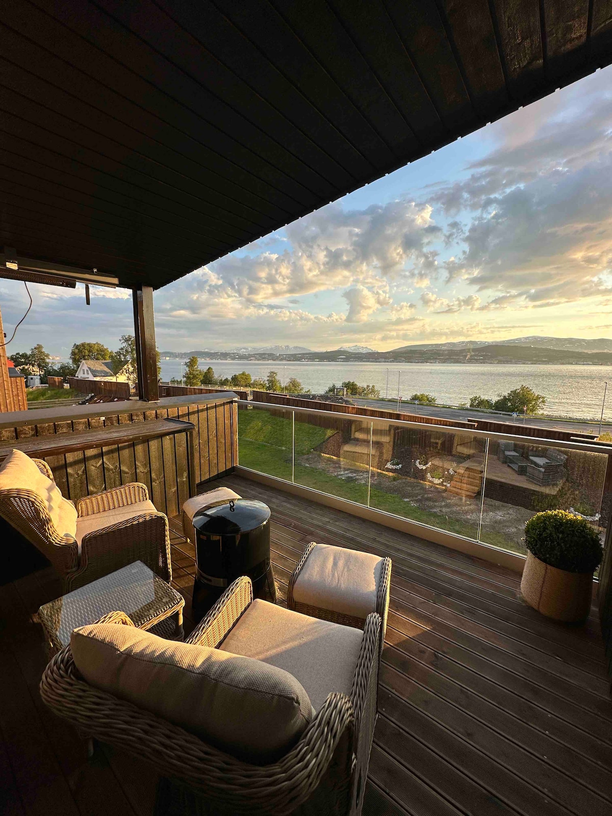 A furnished balcony is shown, featuring comfortable wicker seating with soft cushions. A small table stands nearby, providing a space for drinks. The view includes a wide expanse of water and a sky filled with clouds, illuminated by soft sunset colors.