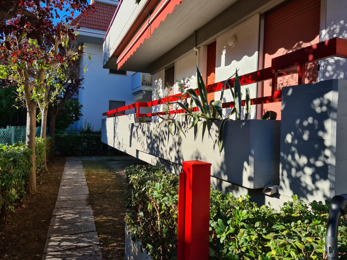 A sunlit walkway leads to the elevated terrace of the apartment, framed by vibrant red railings. Lush greenery and potted plants adorn the terrace, contributing to the inviting appearance of the exterior. The building is surrounded by trees, providing gentle shade.