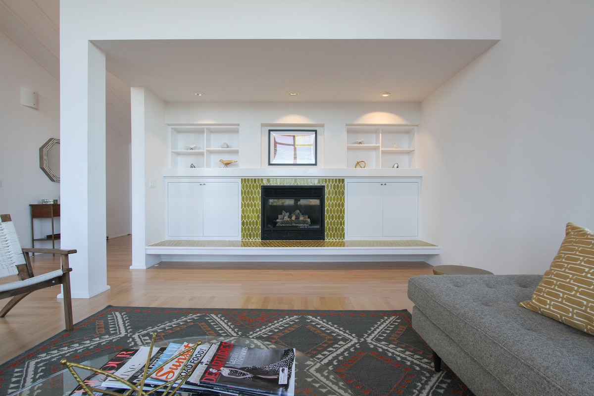 A minimalist living area features a modern fireplace framed by built-in shelves, displaying decorative items. A gray sectional sofa is placed nearby, complemented by a patterned area rug. Natural light enters through large windows, enhancing the clean and spacious design.
